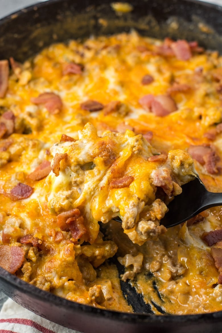 Close up of a spoonful of cheeseburger casserole being pulled out of a pan, topped with bacon bits and melted cheese.