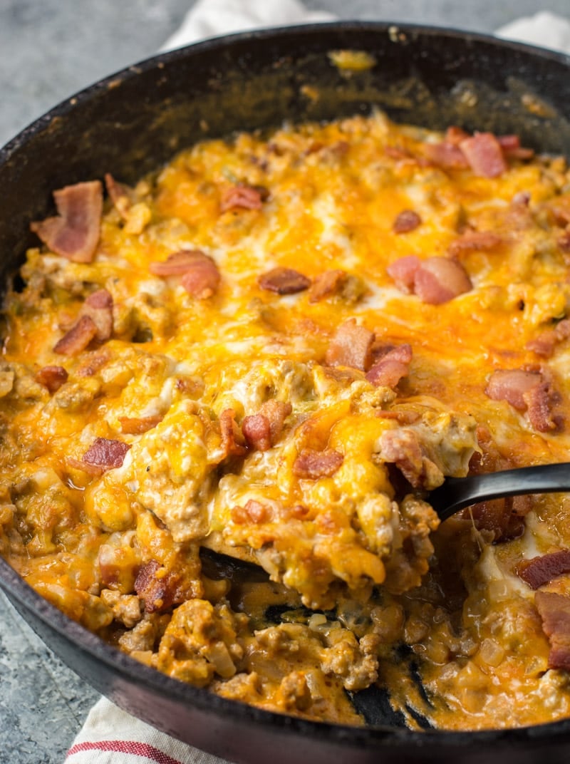 A cheeseburger casserole, topped with melted cheese and pieces of bacon, in a skillet with a serving spoon removing a spoonful. 