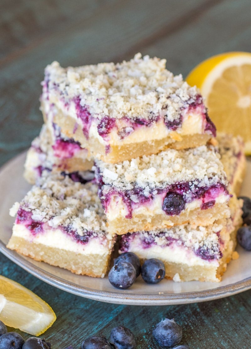 A stack of blueberry cheesecake bars on a plate, surrounded by fresh blueberries and lemon slices