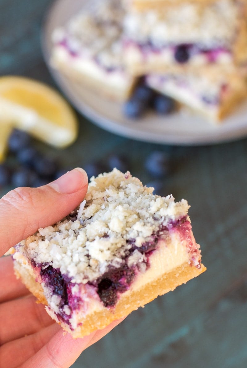 A hand holding a blueberry cheesecake bar, with more bars, blueberries, and lemon slices in the background