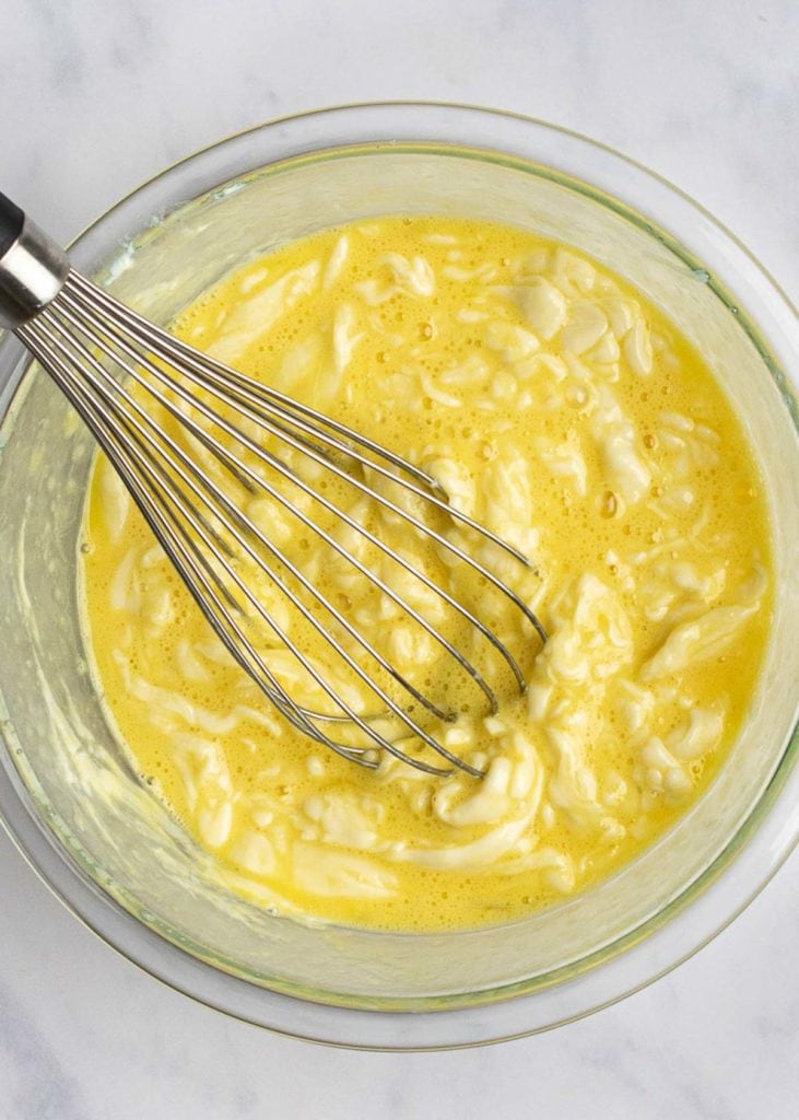 A whisk whisking eggs into melted cheese in a mixing bowl