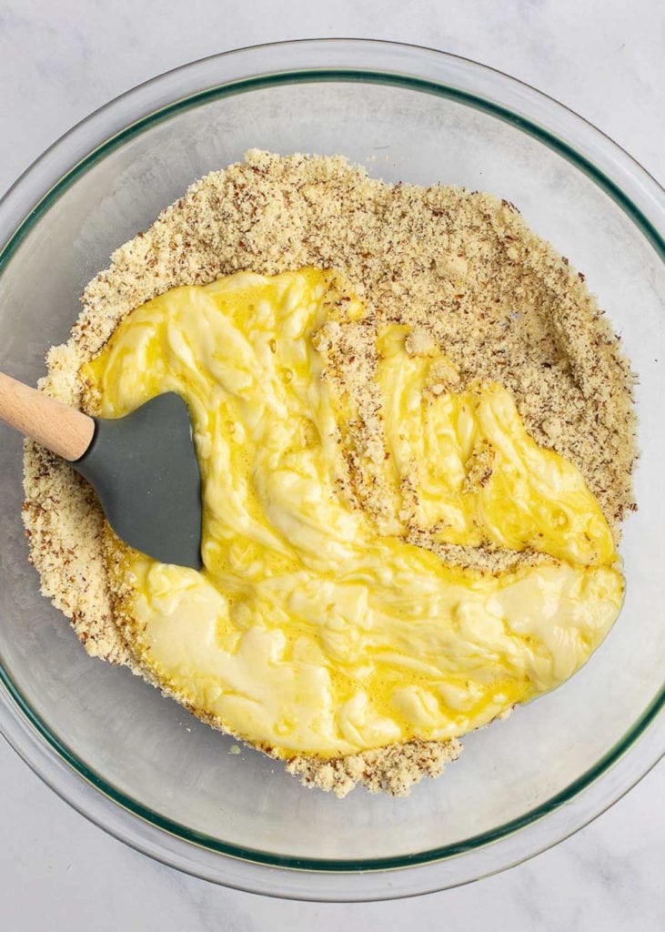 A mixing bowl with dry ingredients topped with an egg and melted cheese mixture, with a rubber spatula stirring it