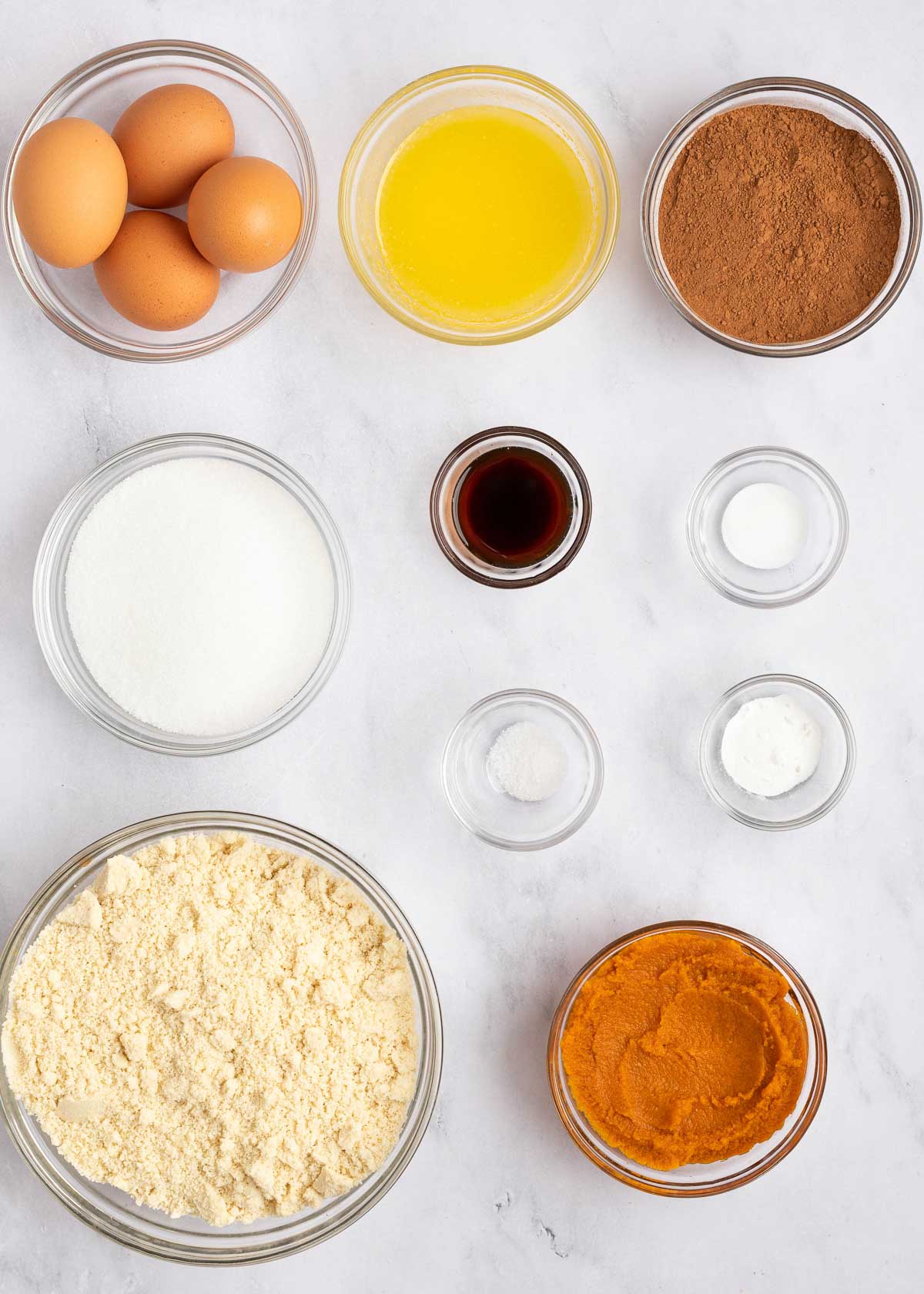 Overhead view of the ingredients needed for keto chocolate cake: a bowl of eggs, a bowl of melted butter, a bowl of cocoa powder, a bowl of almond flour, a bowl of pumpkin puree, a bowl of baking soda, a bowl of baking powder, a bowl of salt, a bowl of vanilla extract, and a bowl of monk fruit sweetener