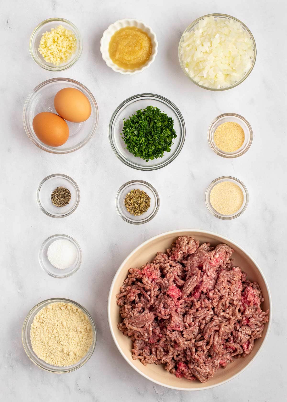 Overhead view of the ingredients needed for the keto meatloaf: a bowl of ground beef, a bowl of onions, a bowl of eggs, a bowl of garlic, a bowl of applesauce, a bowl of onion powder, a bowl of garlic powder, a bowl of ground oregano, a bowl of salt, a bowl of pepper, and a bowl of almond flour
