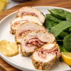 Five slices of chicken cordon bleu on a plate with spinach, mustard, and a lemon slice, with more slices in the background