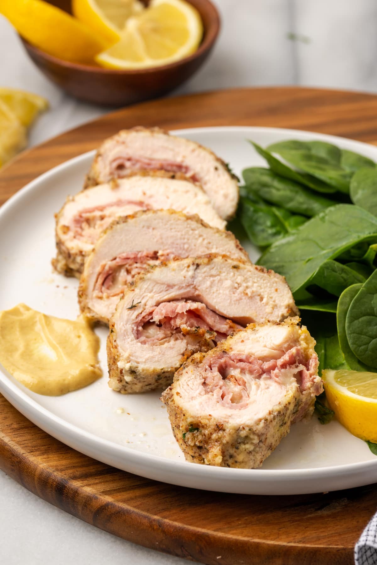 Five slices of chicken cordon bleu on a plate with spinach, mustard, and a lemon slice, with more slices in the background