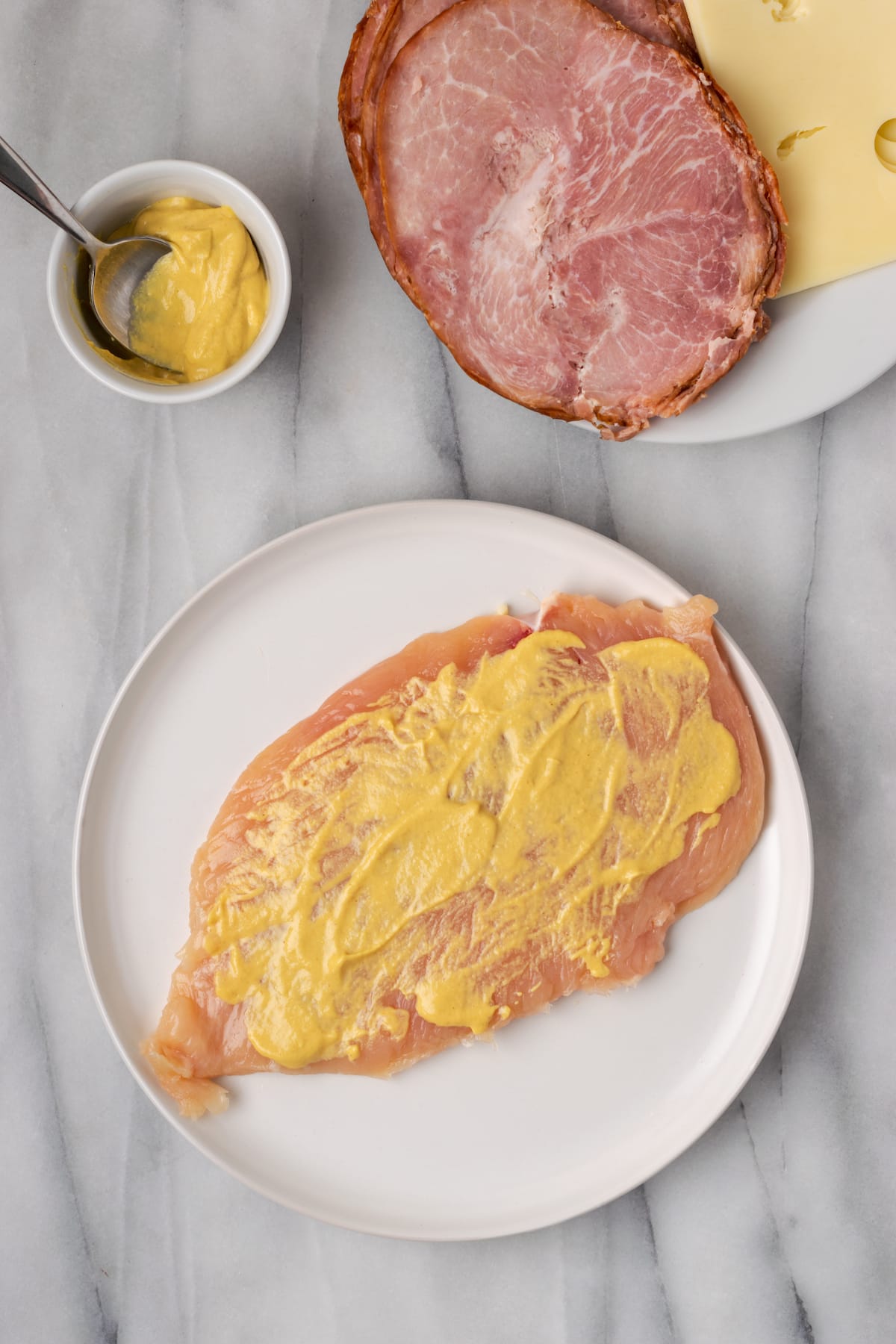 Overhead view of a chicken breast on a plate, covered in mustard, next to a plate of ham and cheese and a ramekin of mustard