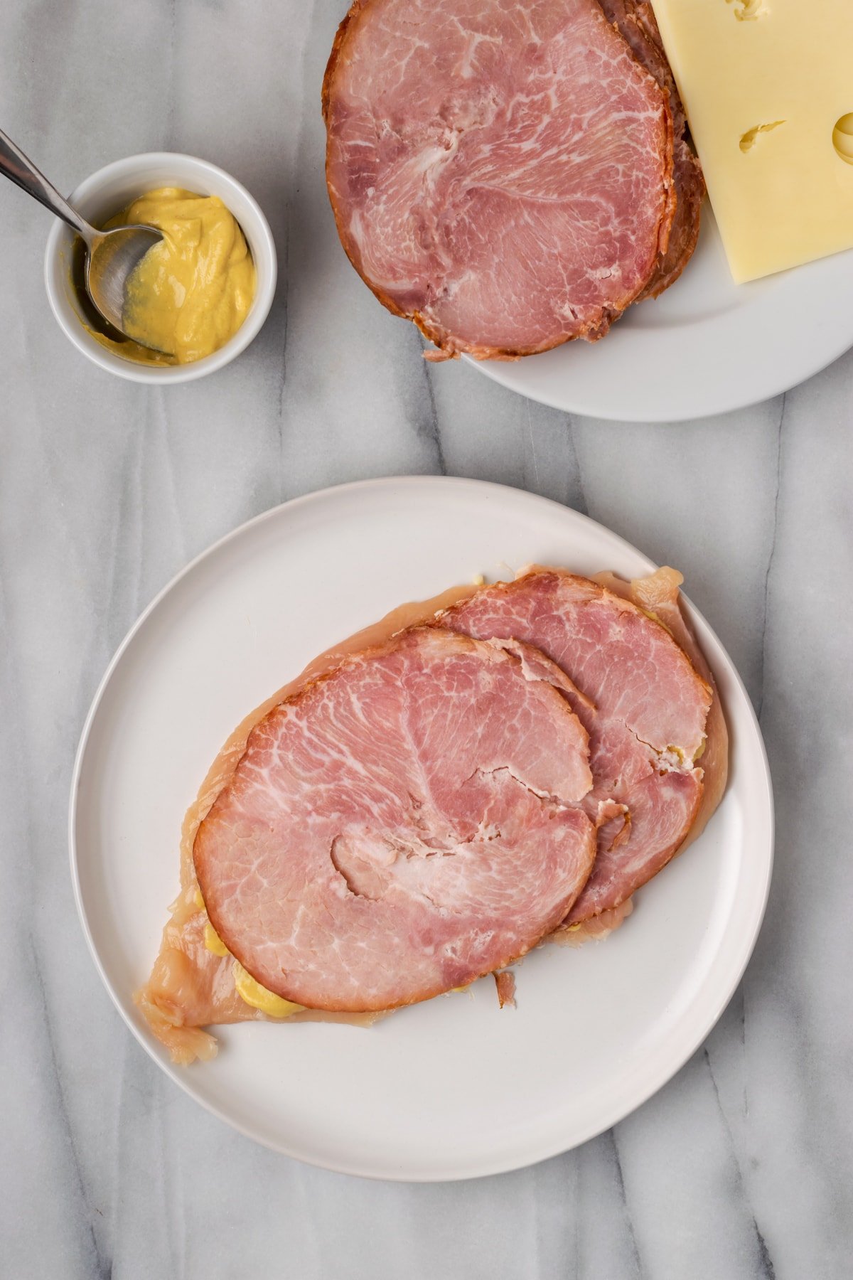 Overhead view of two ham slices on top of a chicken breast on a plate, next to a plate of ham and cheese and a ramekin of tmustard