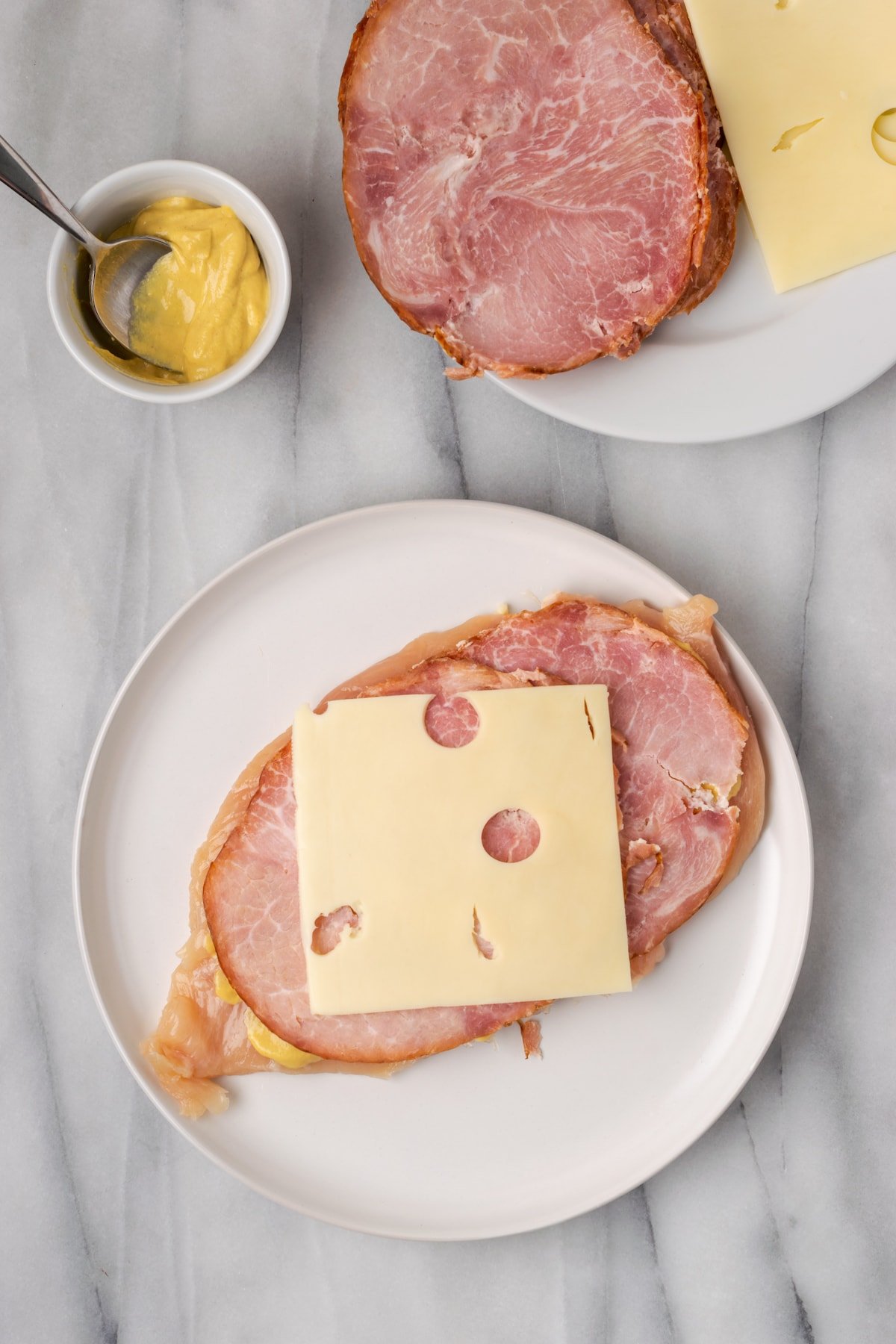 Overhead view of a chicken breast on a plate, topped with hap and Swiss cheese, next to a plate of ham and cheese and a bowl of mustard