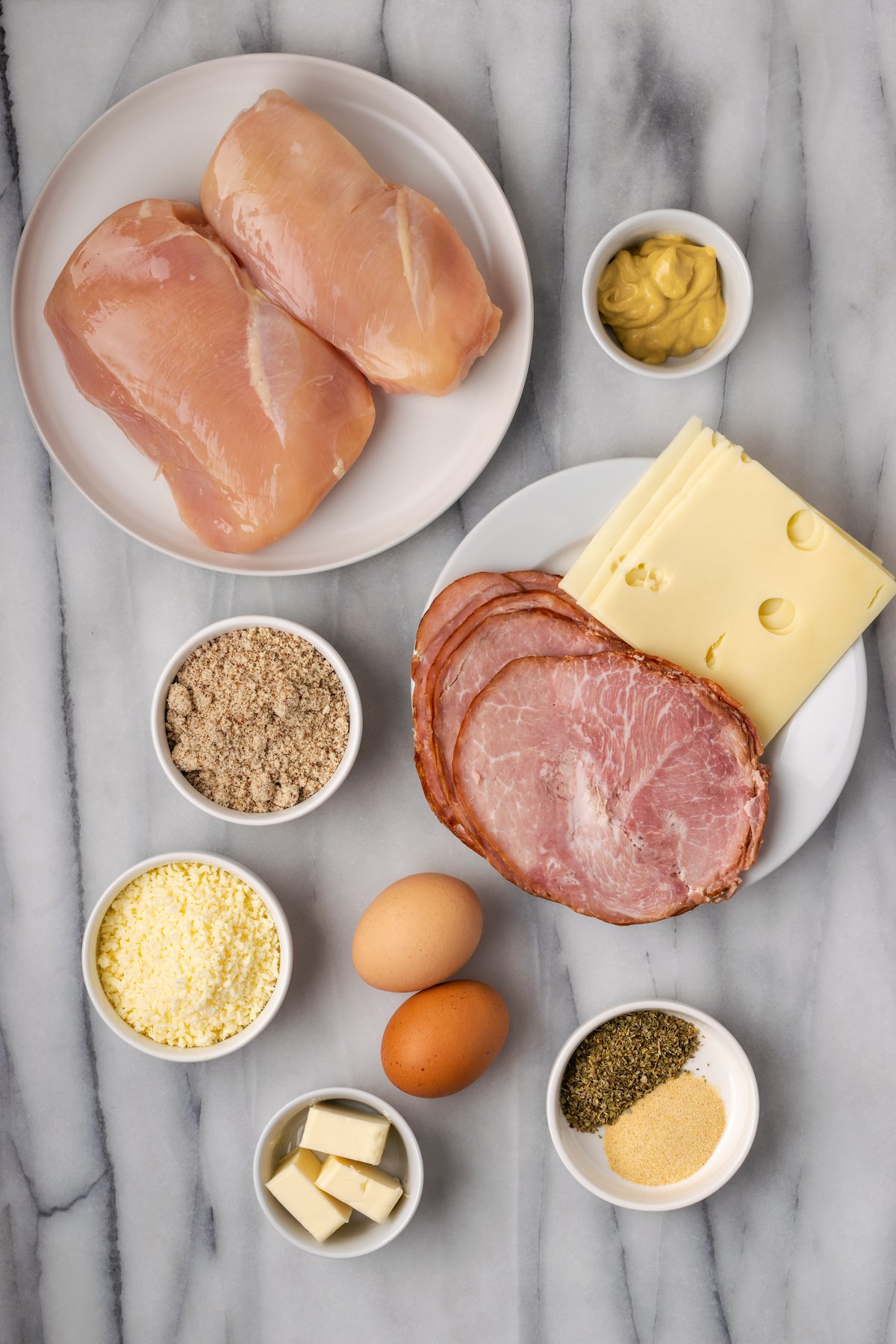 Overhead view of the ingredients needed for chicken cordon bleu: a plate of chicken breasts, a plate with ham and cheese, a bowl of dijon, a bowl of almond flour, a bowl of parmesan cheese, a bowl of garlic powder and Italian seasoning, a ramekin of butter, and two eggs