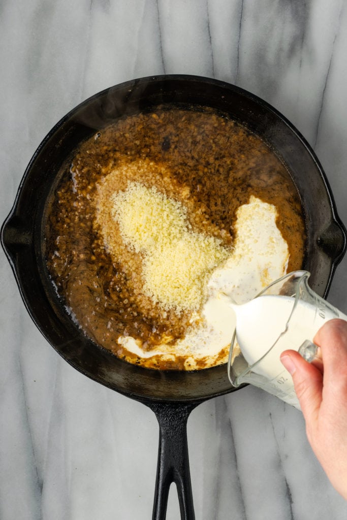 A skillet of wine and garlic, with a pile of parmesan cheese on top, with a hand pouring a pyrex of cream into it