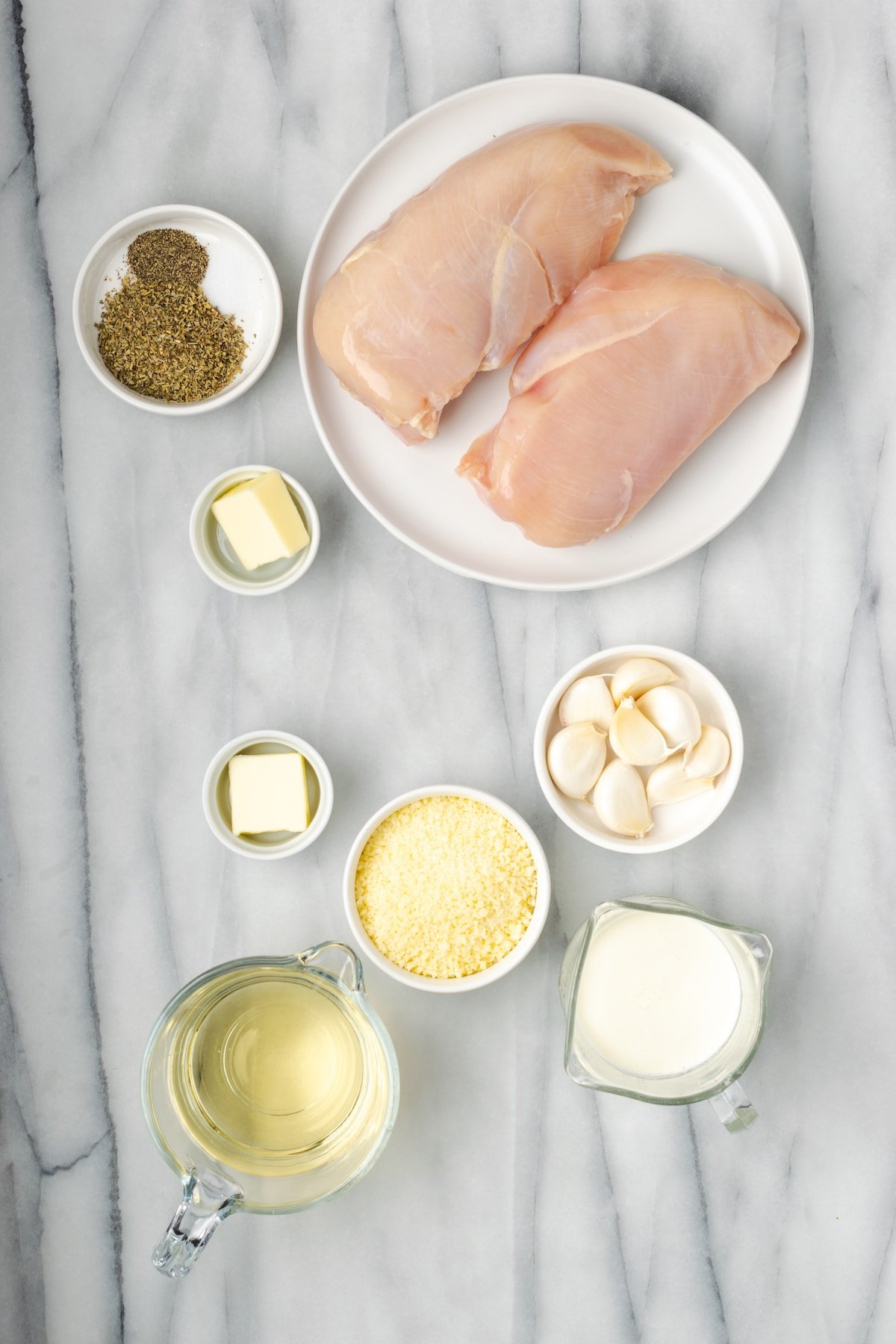 Overhead view of the ingredients needed for garlic parmesan chicken: two chicken breasts, a bowl of garlic, a bowl of parmesan cheese, a glass of white wine, a glass of heavy cream, two ramekins of butter, and a bowl with salt, pepper, and Italian seasoning