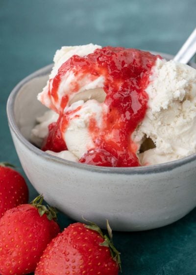 Close up of a bowl of vanilla ice cream topped with strawberry sauce, with a spoon in it, and some strawberries