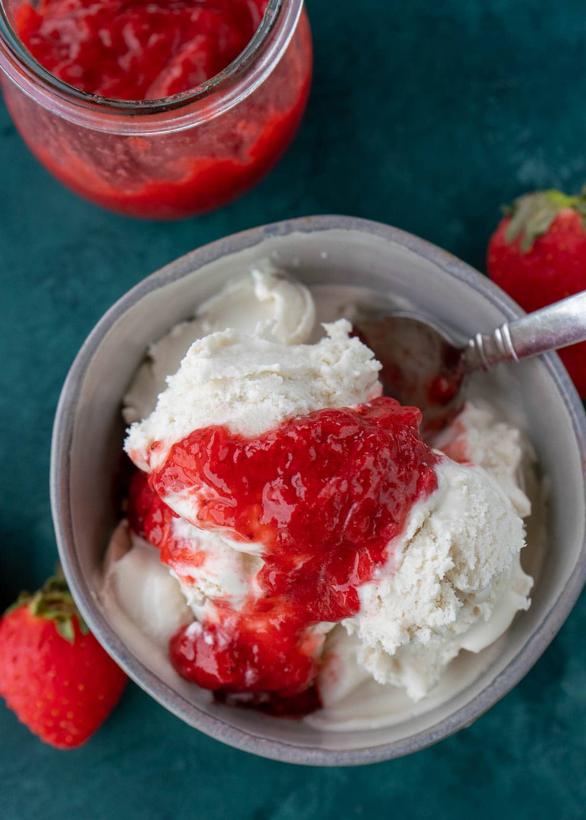 Overhead view of a bowl of vanilla ice cream topped with strawberry sauce, with a spoon in it, next to some strawberries and a jar of sauce