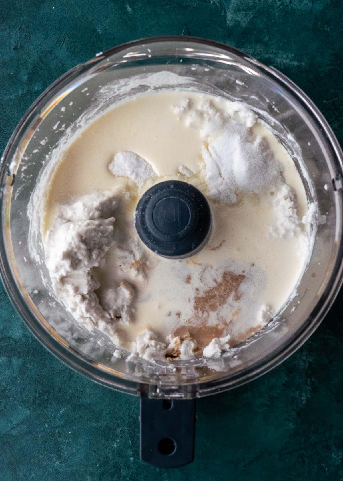 Overhead view of coconut cream, heavy cream, sweetener, salt, and vanilla in a food processor