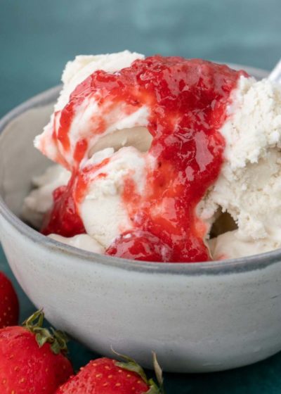 Close up of a bowl of vanilla ice cream topped with strawberry sauce, with a spoon in it, and some strawberries