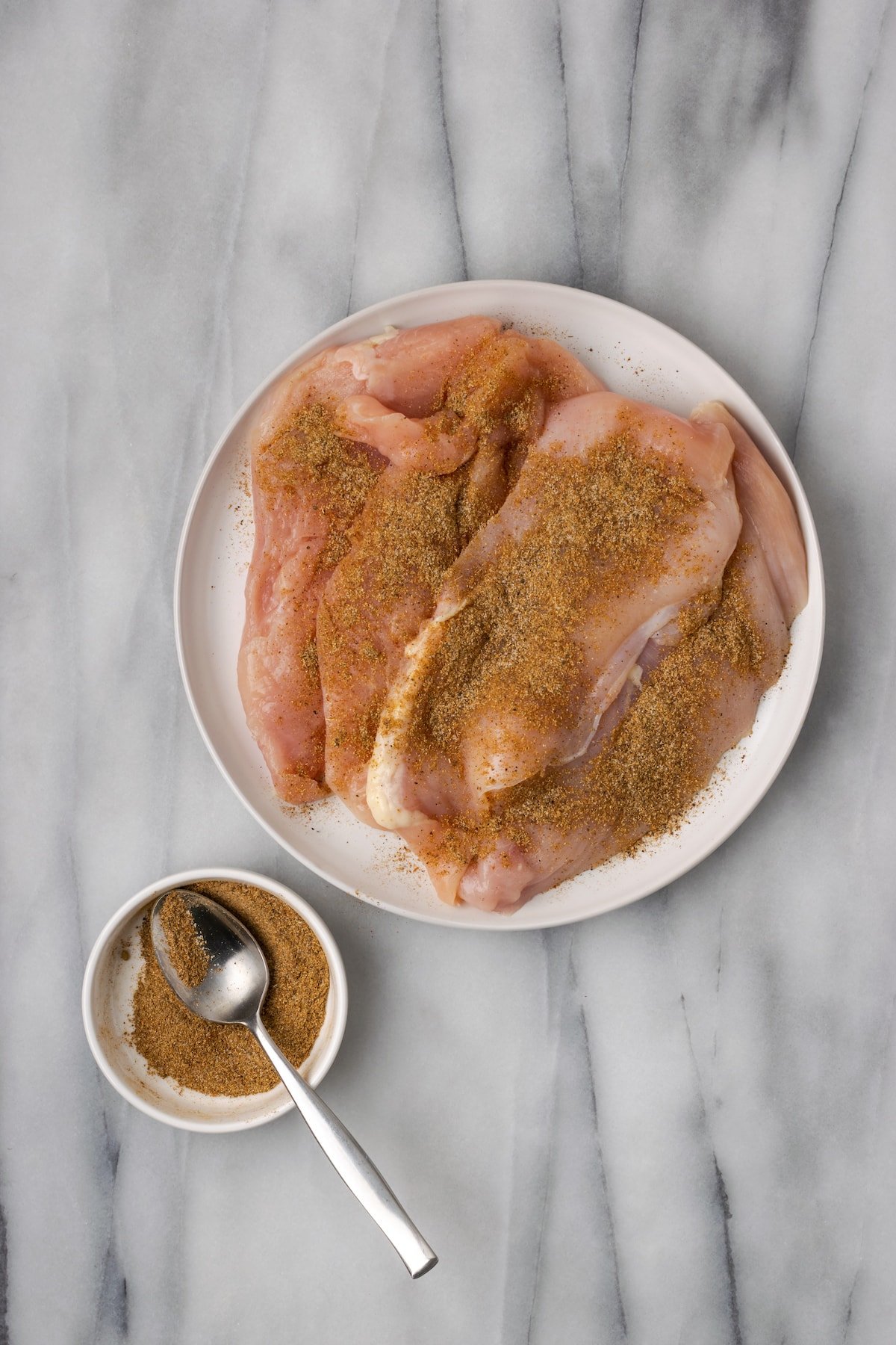 Overhead view of four seasoned, raw chicken breasts on a plate, next to a bowl of spice rub with a spoon in it