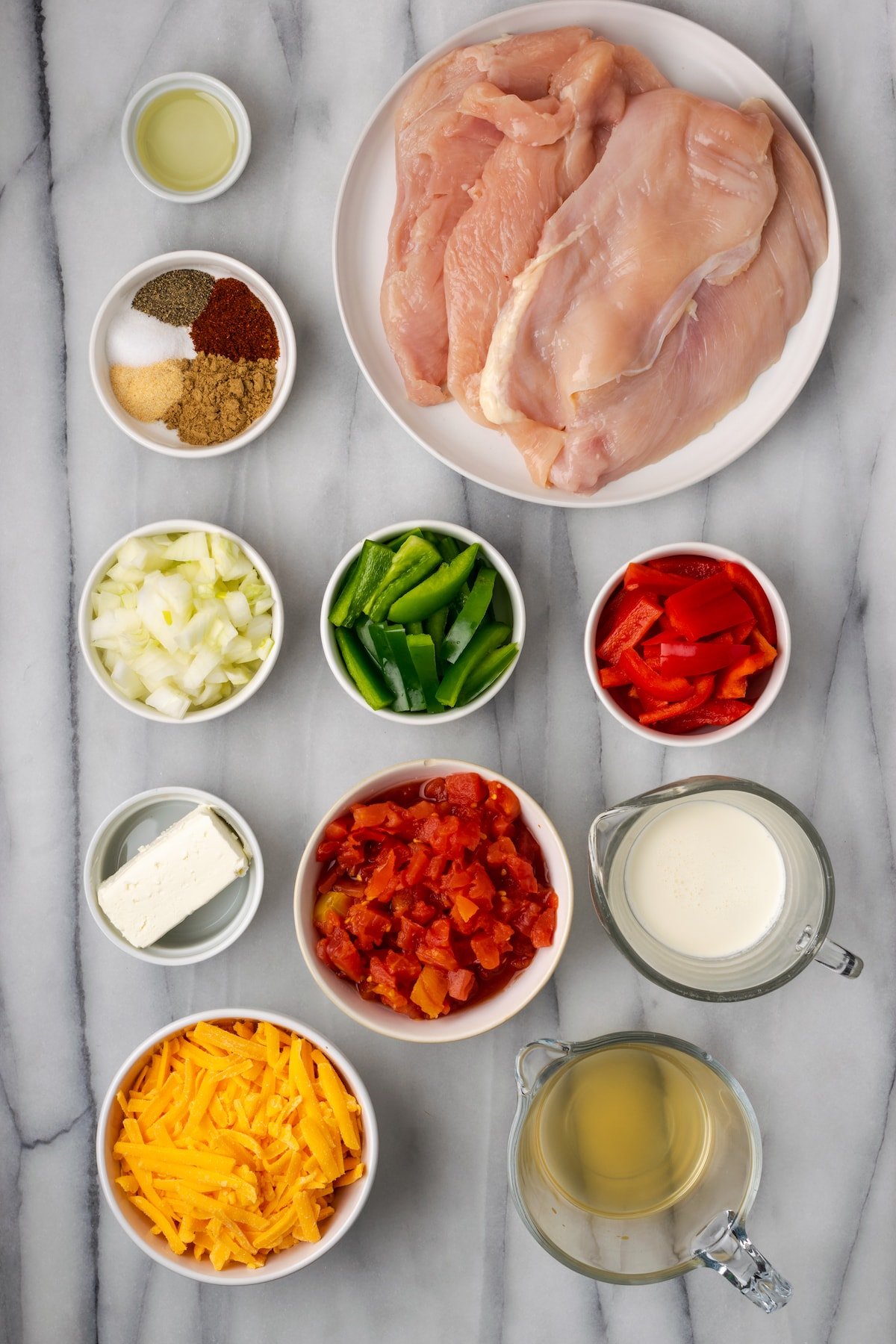 Overhead view of the ingredients needed for Santa Fe chicken: a plate of chicken breasts, a bowl of olive oil, a bowl of salt, pepper, cumin, chili powder, and garlic powder, a bowl of onions, a bowl of green bell peppers, a bowl of red bell peppers, a bowl of cream cheese, a bowl of Ro-tel, a bowl of shredded cheddar cheese, a pyrex of heavy cream, and a pyrex of chicken broth