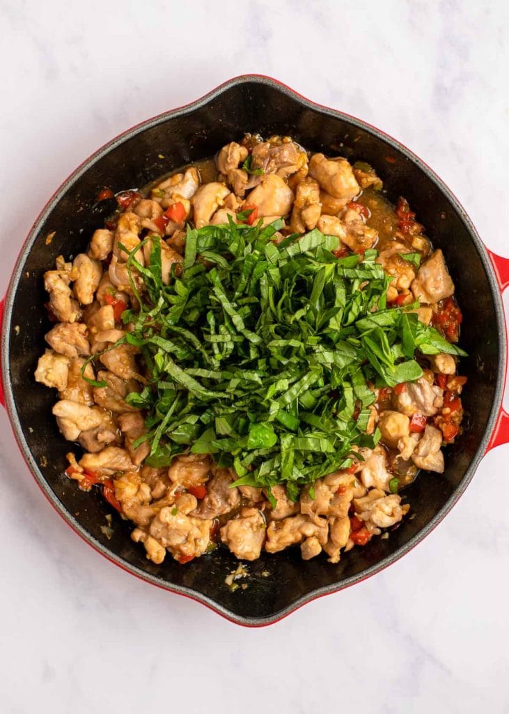 thai basil added to a skillet full of saucy chicken, peppers, shallots, and garlic