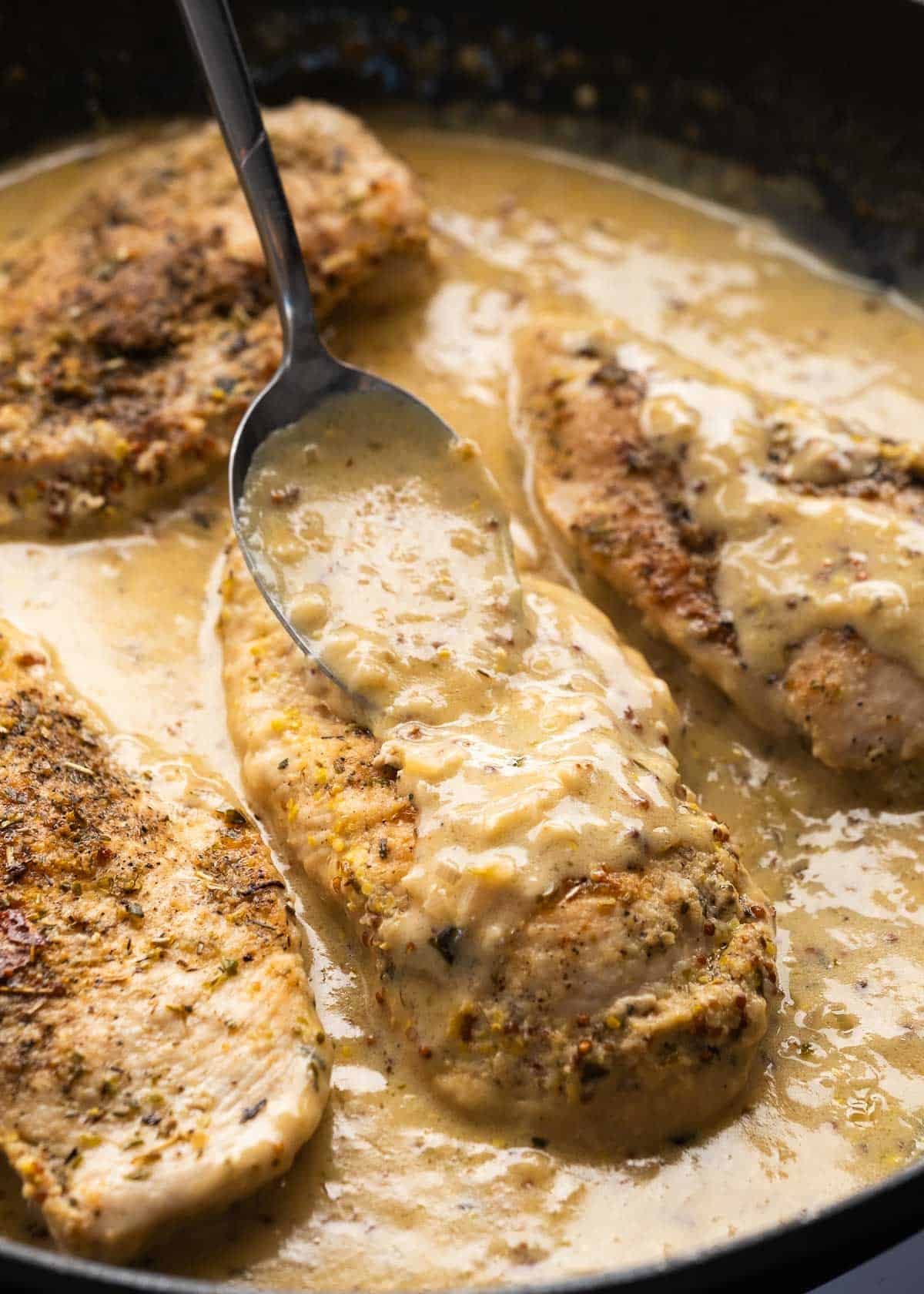 mustard sauce being spooned over chicken breasts in cast iron skillet