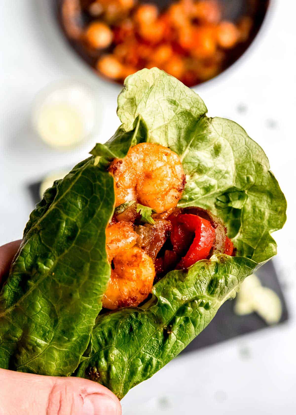 close up of shrimp lettuce wrap being held in hand with ingredients on table in background 
