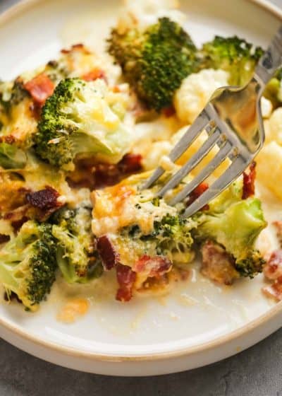 a fork grabbing a bite of cheesy broccoli cauliflower casserole on a white plate