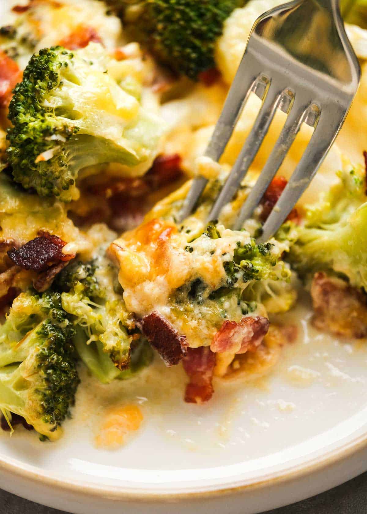 closeup photo of fork picking up a cheesy bite of broccoli cauliflower casserole on a white plate