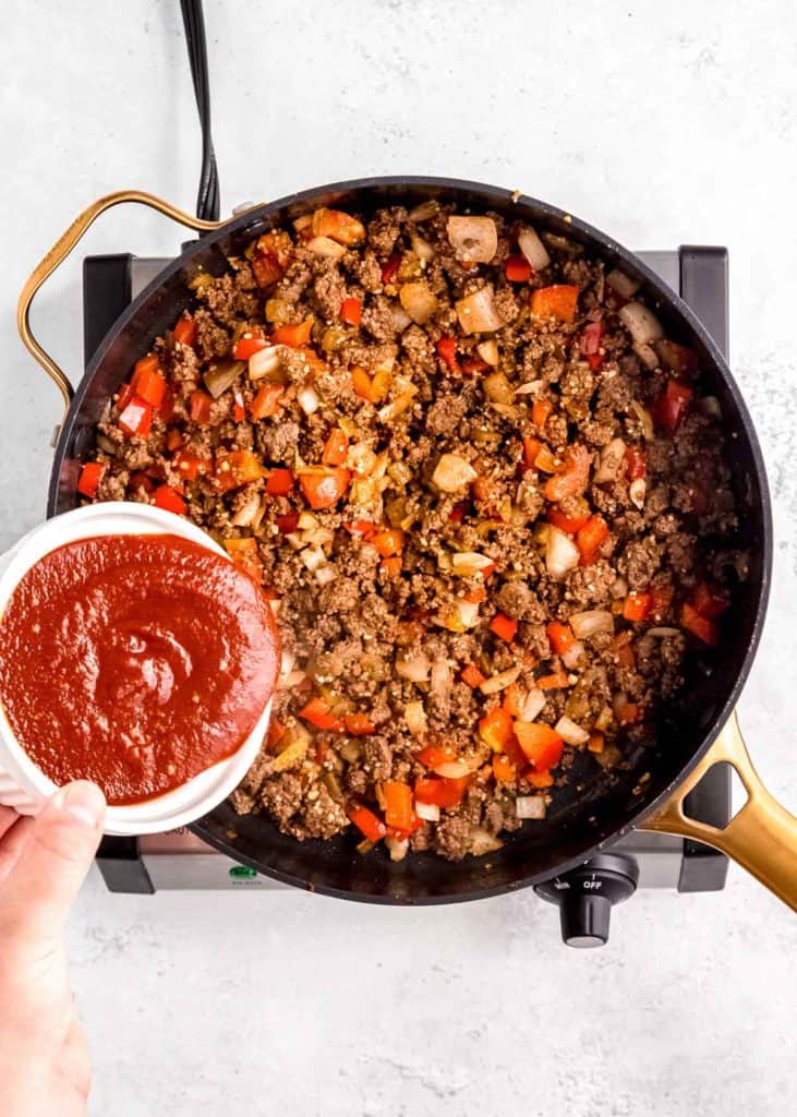 a hand pouring enchilada sauce into the beef and veggies in a skillet