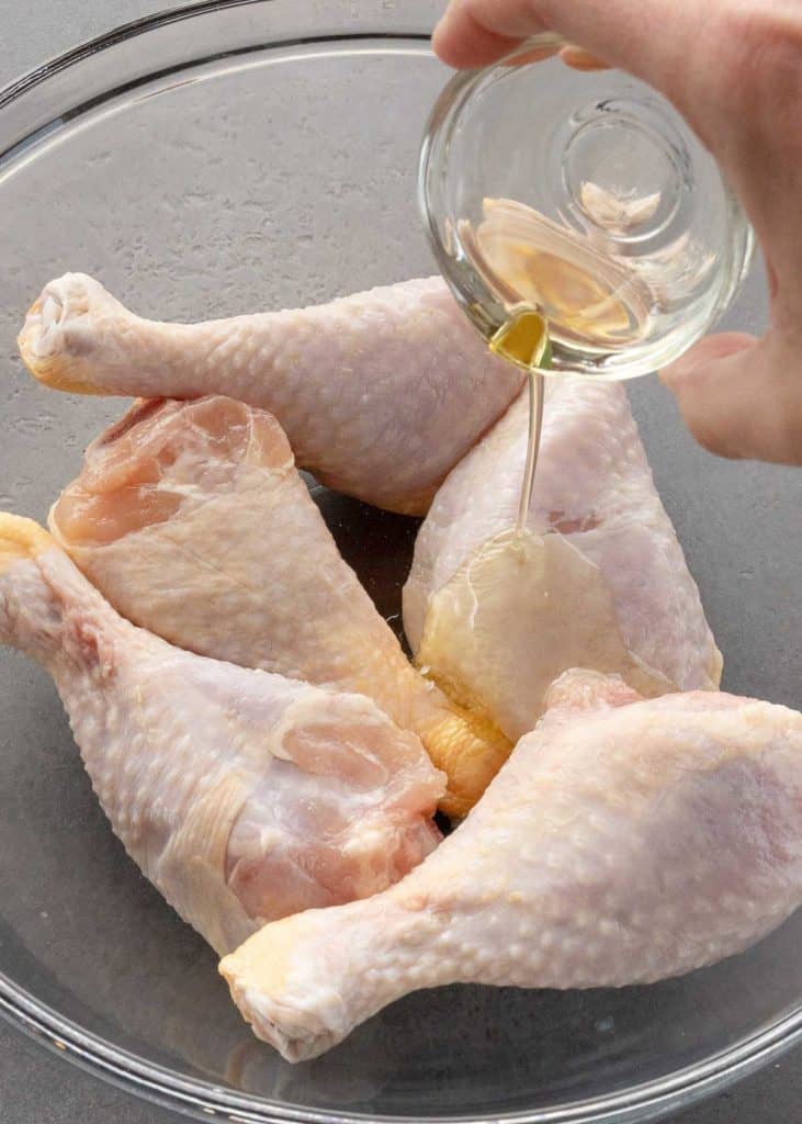 olive oil being poured on chicken legs in a glass bowl