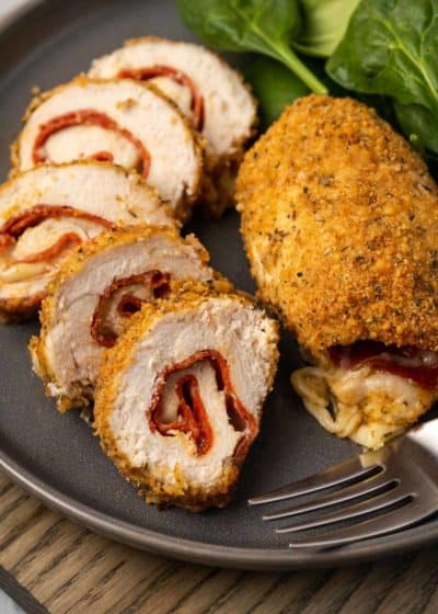 slices of juicy pepperoni stuffed chicken beside a whole breaded stuffed chicken breast on a black plate with a salad on the side