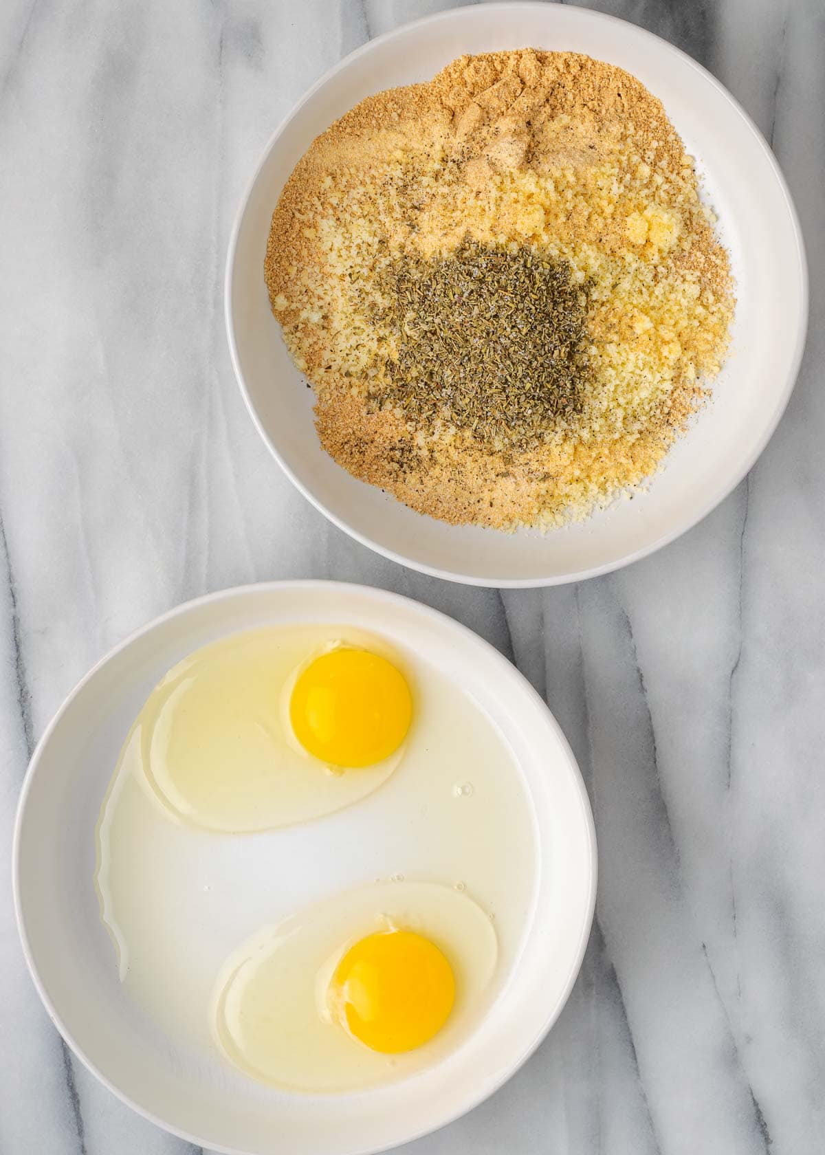 a breading station with two shallow white bowls. One is filled with the egg wash, and the other is filled with the ground pork rinds, parmesan, and seasonings