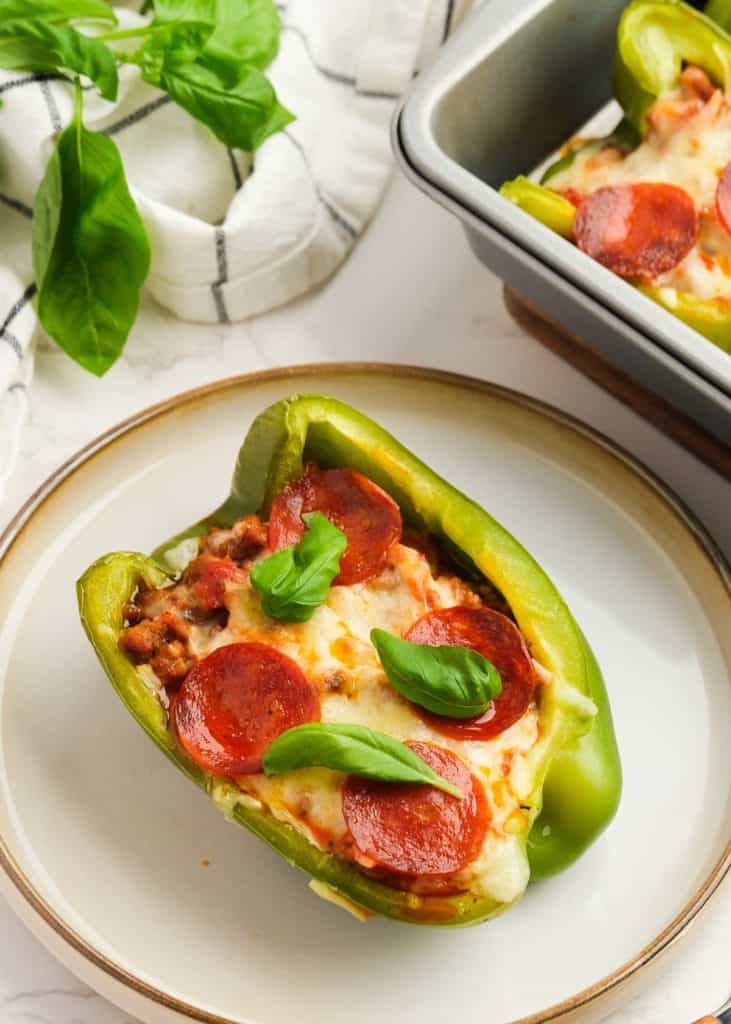 a white plate topped with a pizza stuffed pepper beside a baking dish full of more keto stuffed peppers