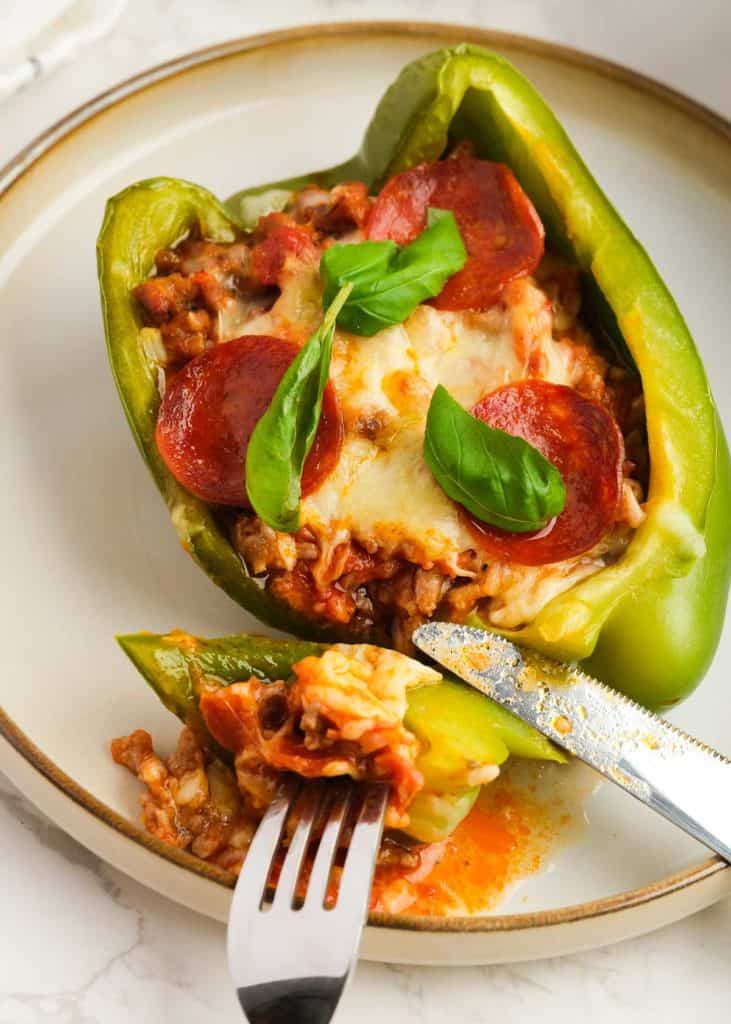 a fork and knife cutting into a pizza stuffed pepper topped with pepperoni and basil on a white plate
