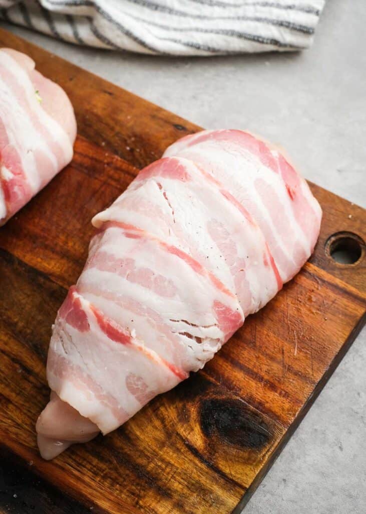 raw bacon wrapped stuffed chicken breast on wooden cutting board