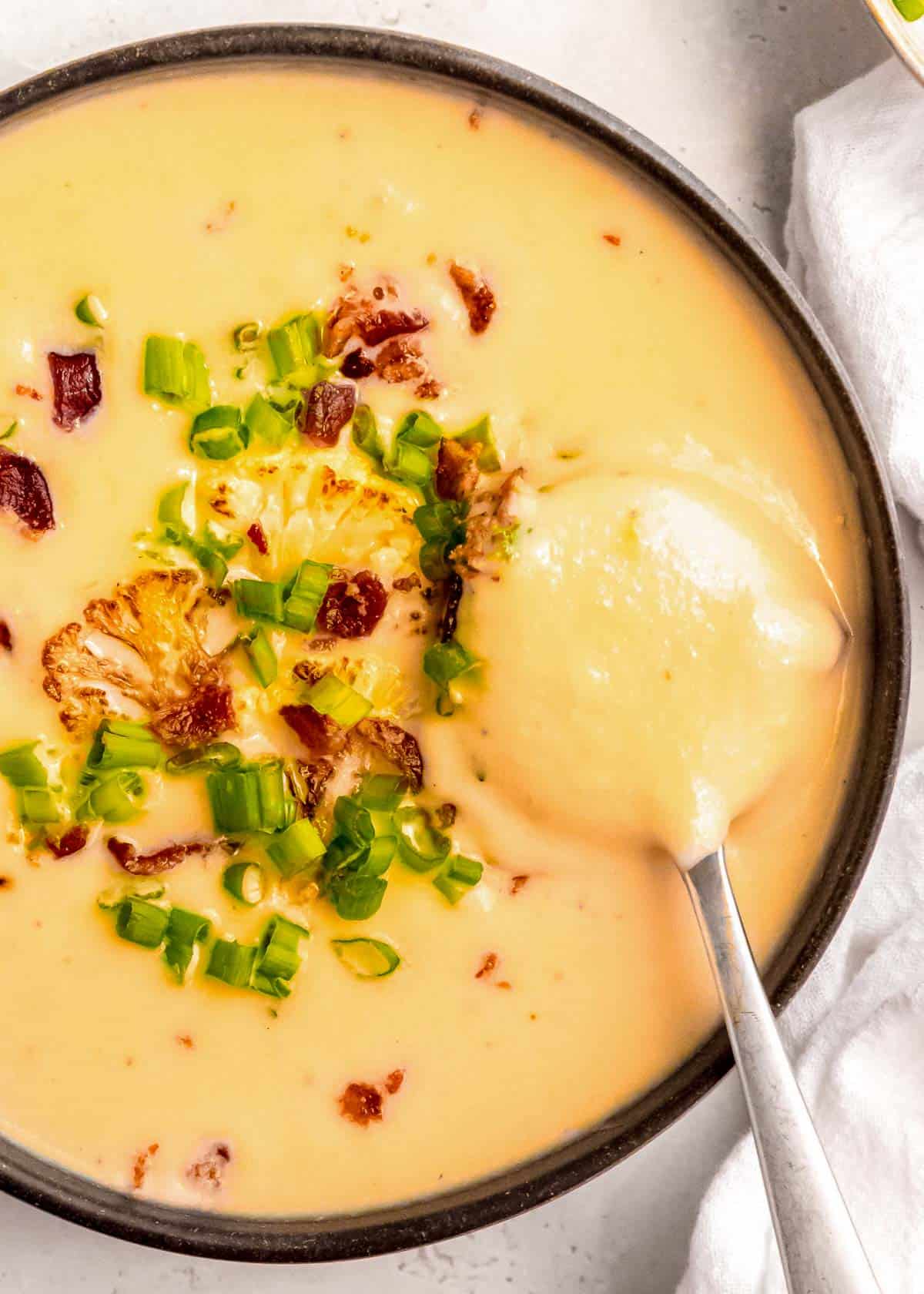 close up overhead image of loaded cauliflower soup in black bowl with silver spoon