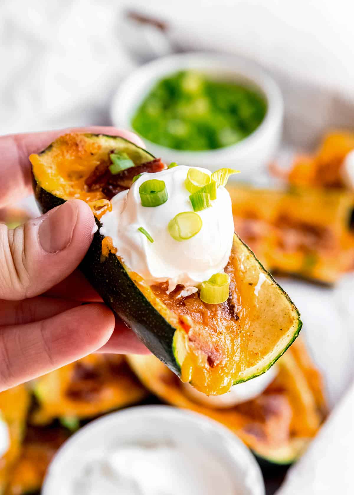 close up image of someone holding a loaded zucchini skin with ingredients in the background