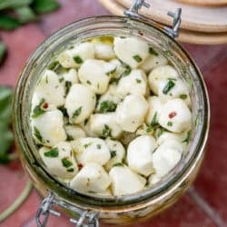 overhead image of marinated mozzarella pearls in a clear jar on a red table with recipe ingredients