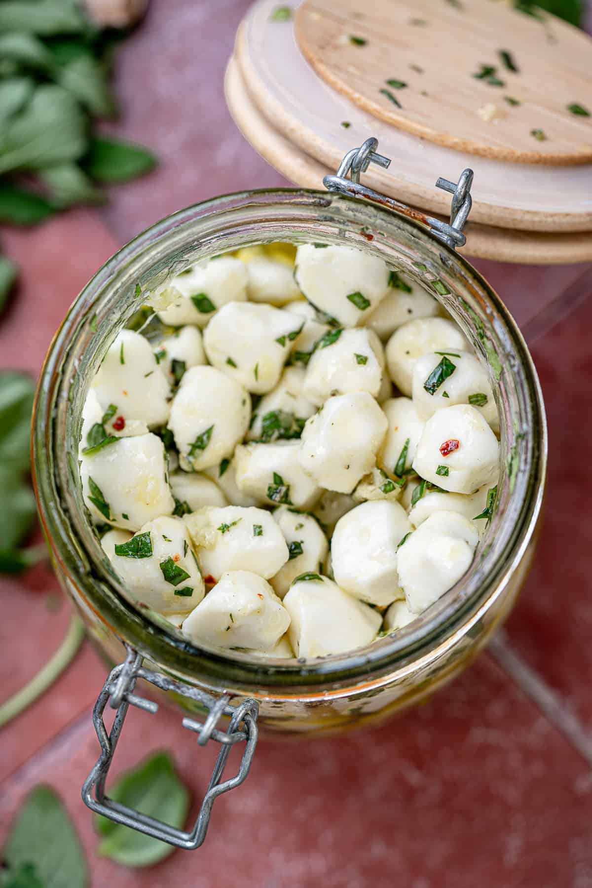 overhead image of marinated mozzarella pearls in a clear jar on a red table with recipe ingredients