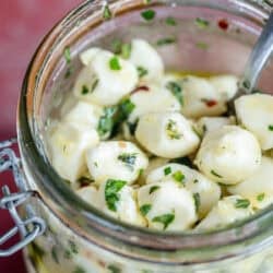 overhead image of marinated mozzarella pearls in a clear jar on a red table with recipe ingredients