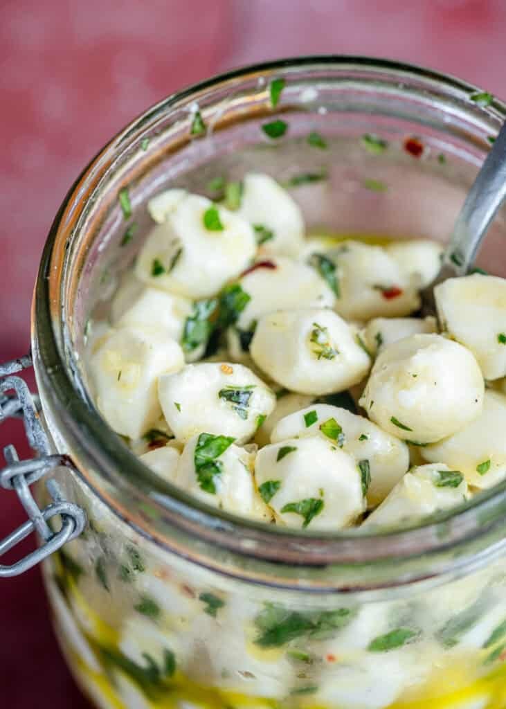 overhead image of marinated mozzarella pearls in a clear jar on a red table with recipe ingredients